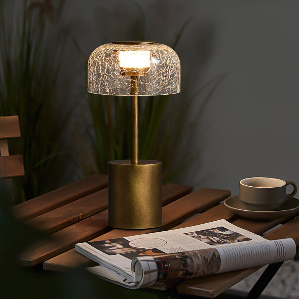 Clérachic crackle glass table lamp with a bronze base, lit, on a slatted wooden outdoor table beside a cup and an open magazine.