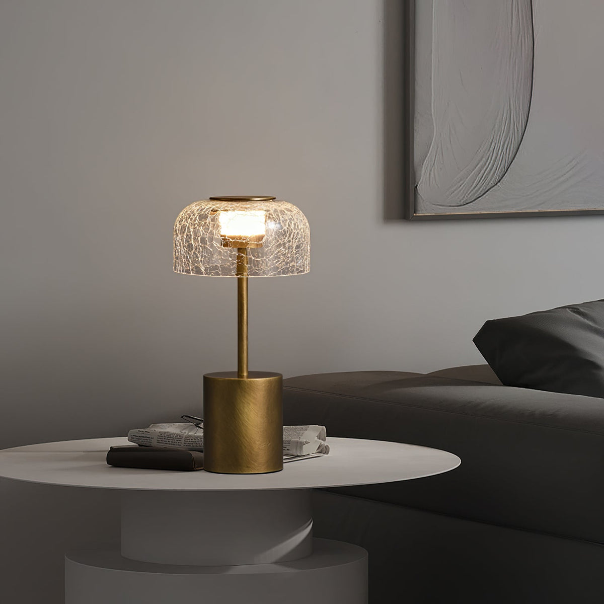 Lit crackle glass table lamp from Clérachic on a round white side table with magazines, in front of a grey sofa and a textured wall panel.