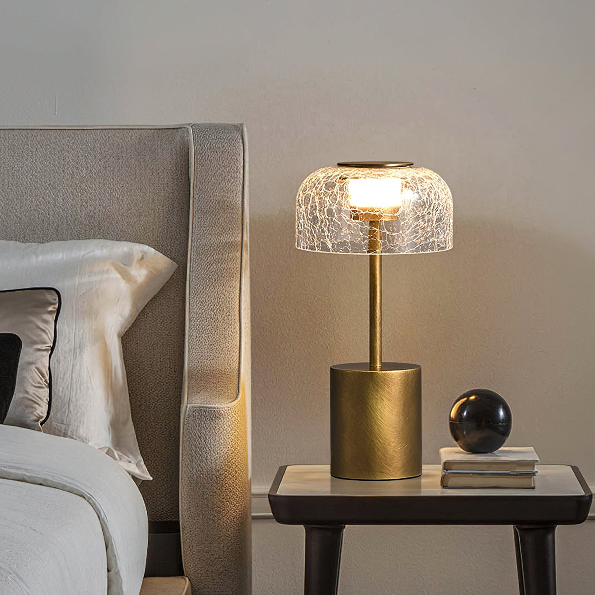 The Clérachic crackle glass lamp on a dark bedside table beside a stack of books and a black sphere, with a pale bed to the left.