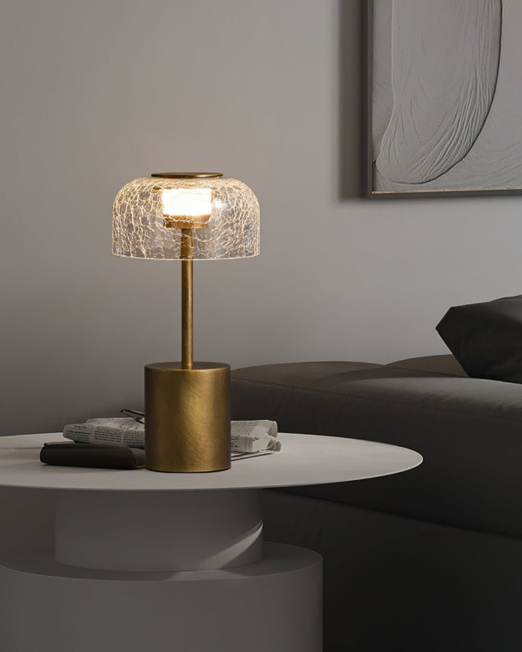 A Clérachic table lamp with a crackle glass dome, lit, standing on a white round table with magazines, beside a dark sofa.