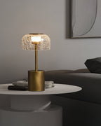 A Clérachic table lamp with a crackle glass dome, lit, standing on a white round table with magazines, beside a dark sofa.