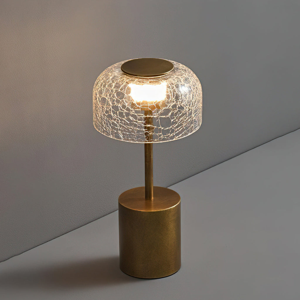 Slightly raised view of this Clérachic table lamp, with a lit crackle glass dome and a brushed bronze round base on a grey surface.