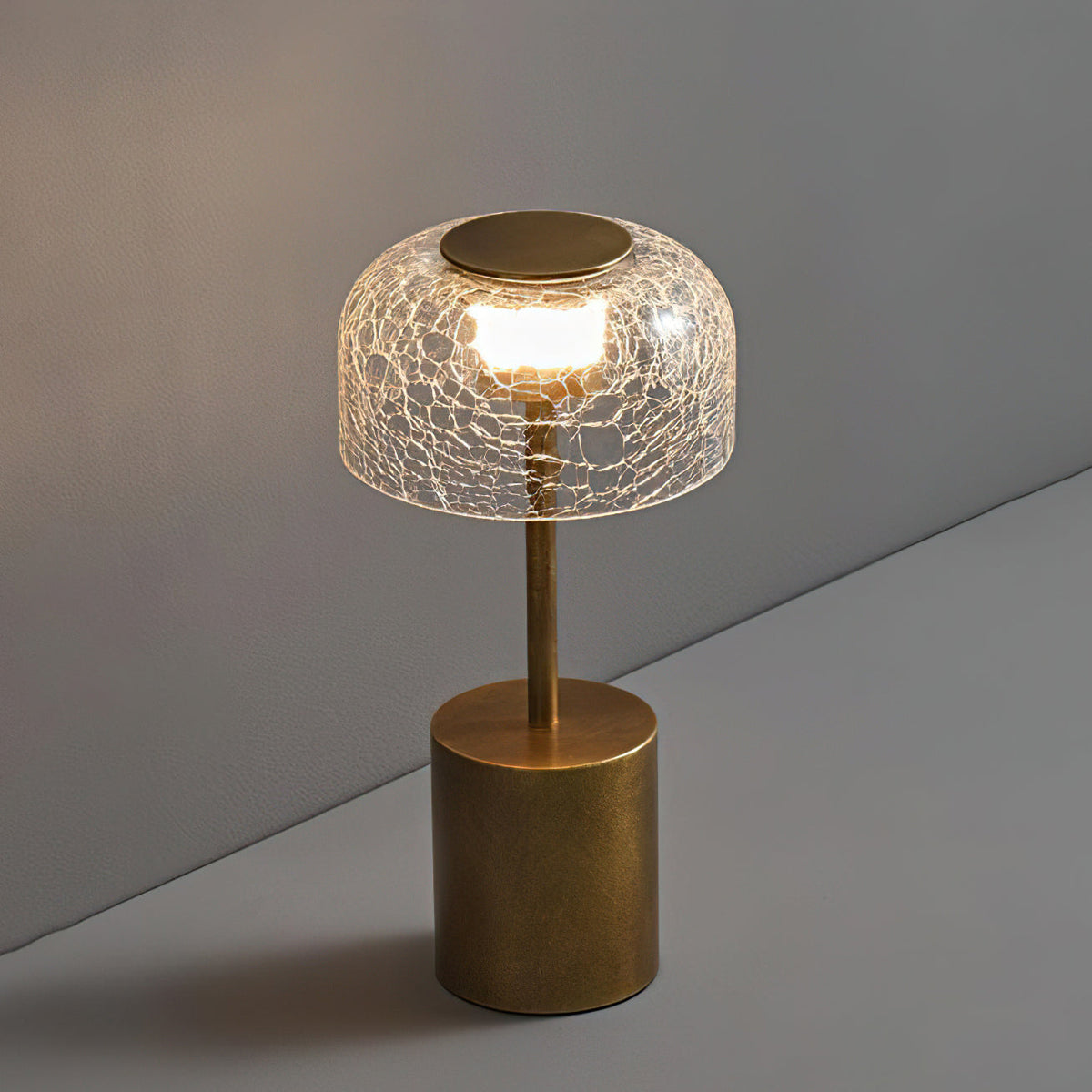 Slightly raised view of this Clérachic table lamp, with a lit crackle glass dome and a brushed bronze round base on a grey surface.