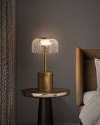 Small Clérachic crackle glass table lamp, lit, on a round side table beside a bed with a tan headboard, in a dim grey room.