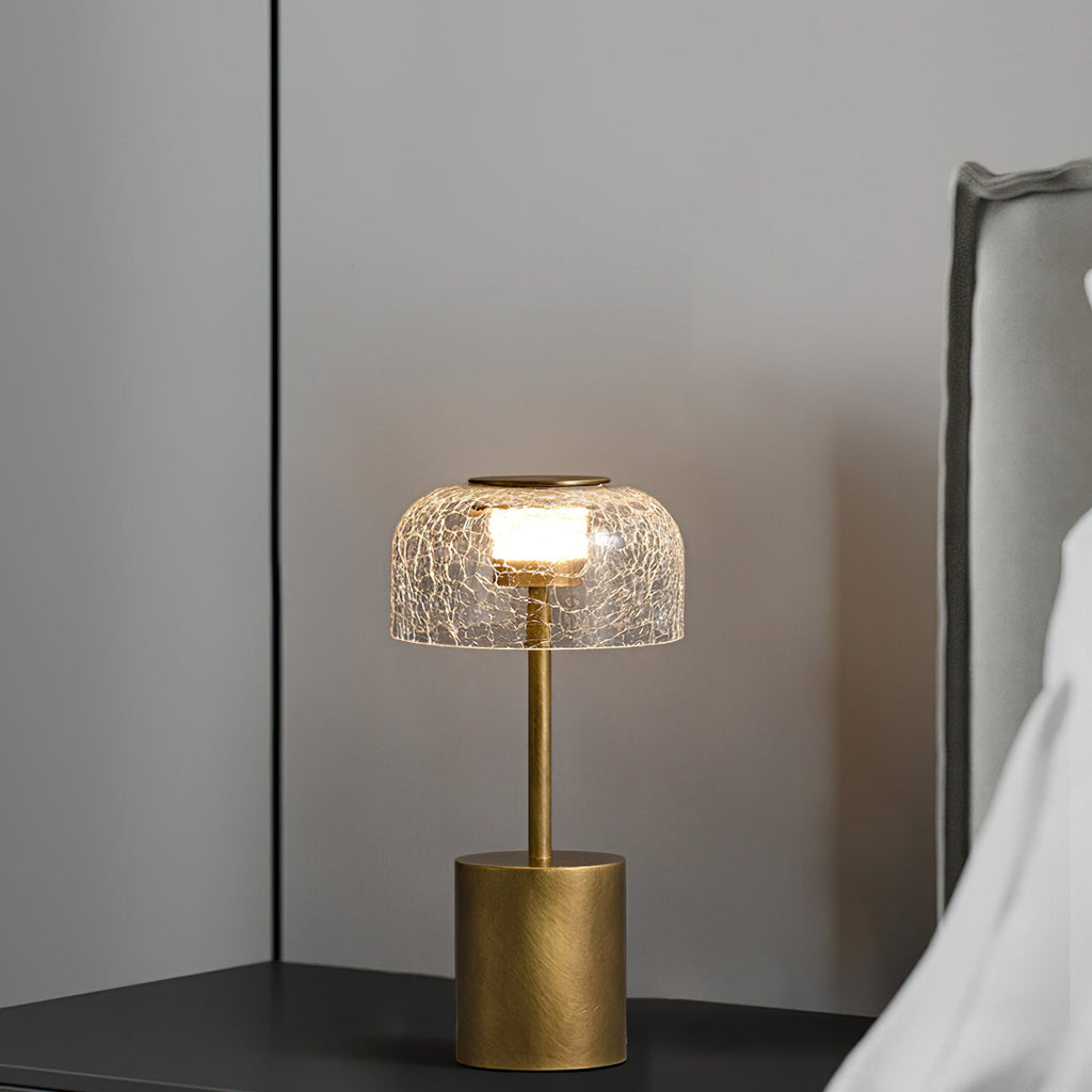 Table lamp with a crackled glass shade from Clérachic, lit, on a dark bedside shelf with a grey headboard and white bedding behind.