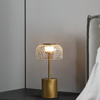 Table lamp with a crackled glass shade from Clérachic, lit, on a dark bedside shelf with a grey headboard and white bedding behind.
