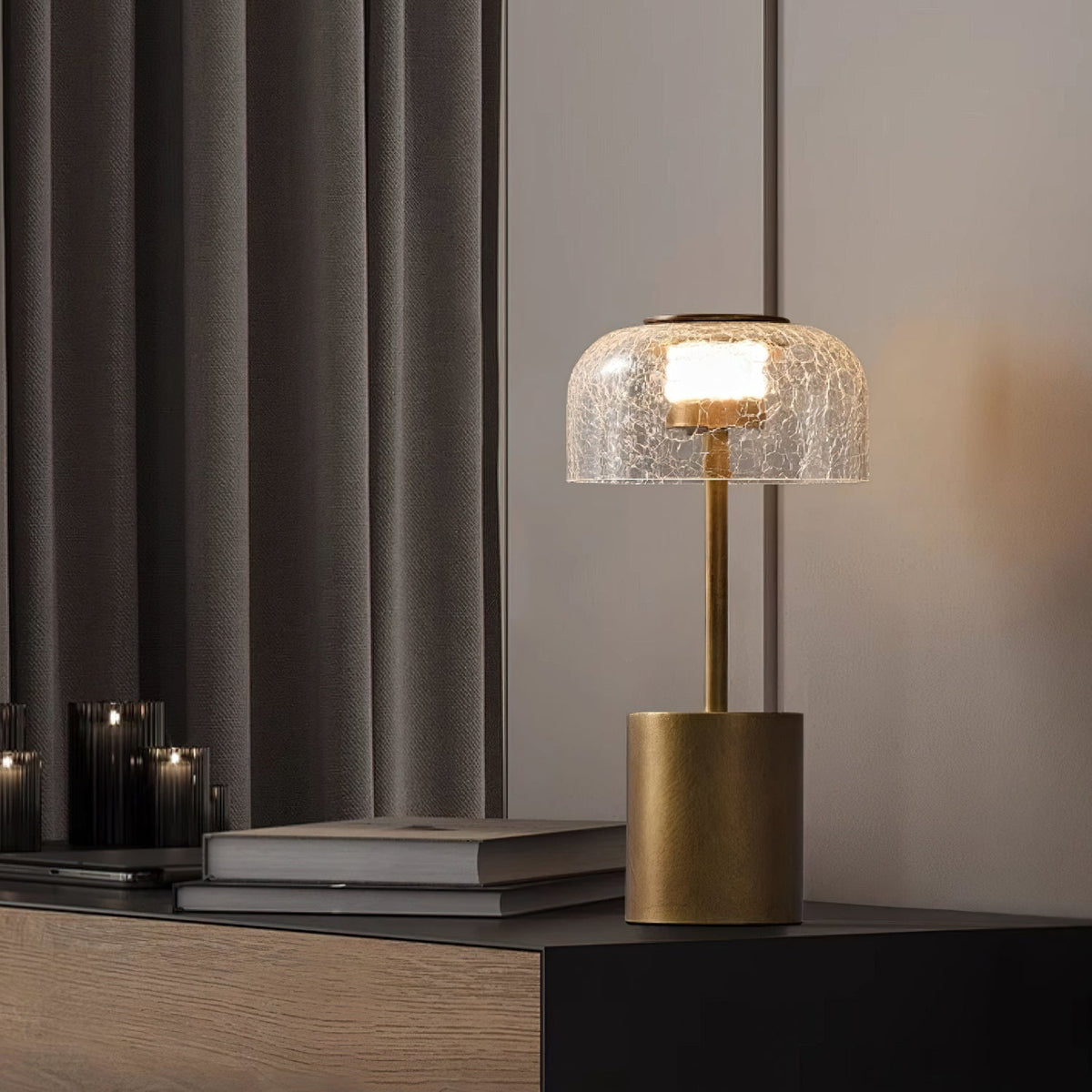 Clérachic table lamp with a lit crackle glass dome and bronze base, on a dark console with books and candles by grey curtains.