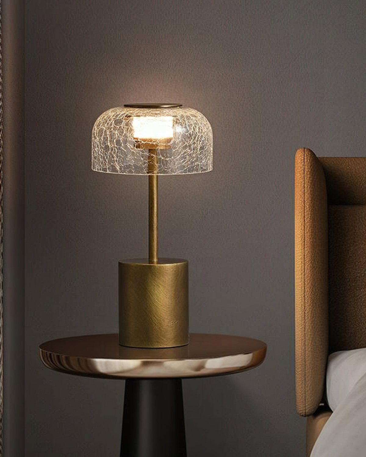 A lit Clérachic crackle glass table lamp on a round side table, with a tan leather headboard and white bedding to the right.