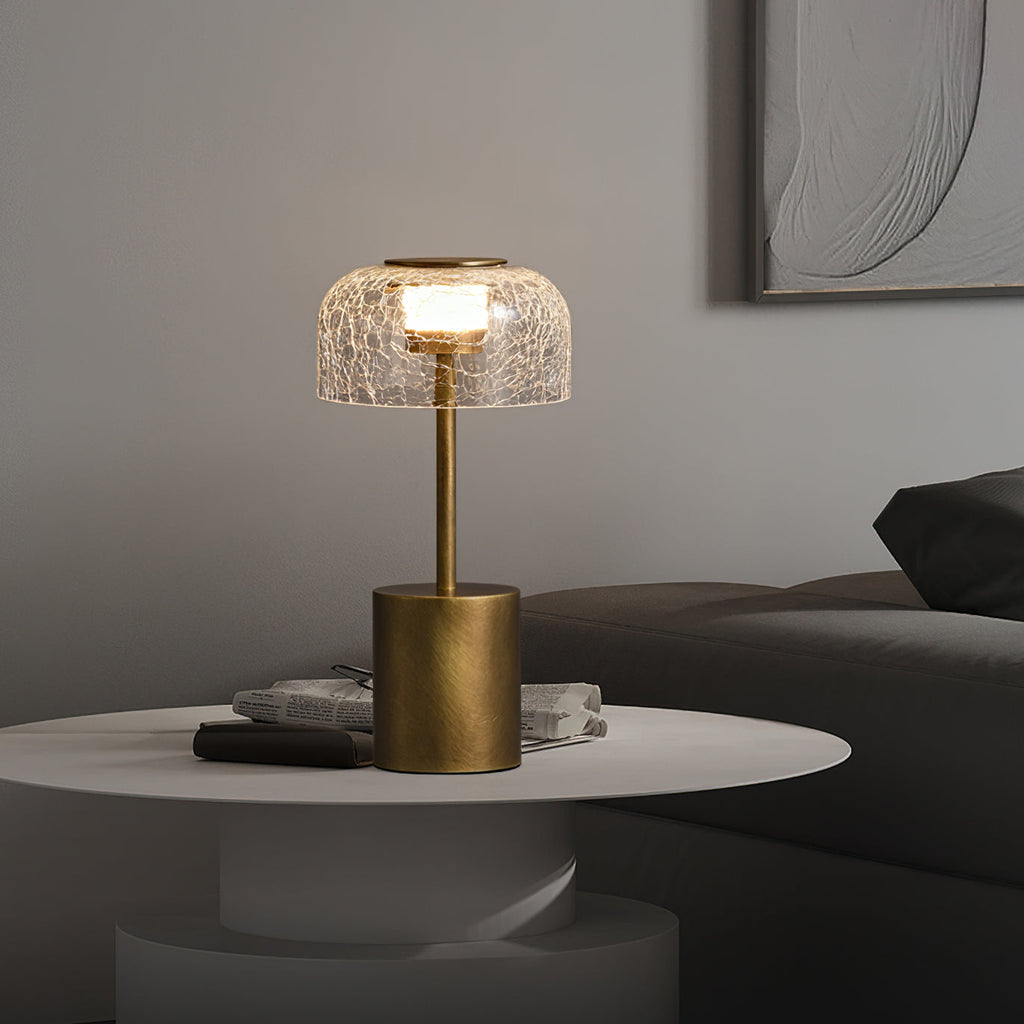 Clérachic crackle glass table lamp lit on a white round table, with a pile of magazines and a dark sofa behind.