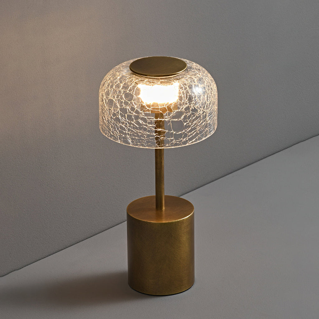 Lit crackle glass table lamp from Clérachic on a grey surface, with a bronze cap on the dome and a diagonal shadow behind.
