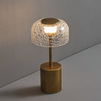 Lit crackle glass table lamp from Clérachic on a grey surface, with a bronze cap on the dome and a diagonal shadow behind.