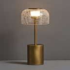 Clérachic crackle glass table lamp with its dome lit warm, standing on a grey surface in front of a grey wall.