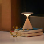 A lit Clérachic table lamp stands on a stack of books, with a dried stem lying across them, beside a wooden cabinet.