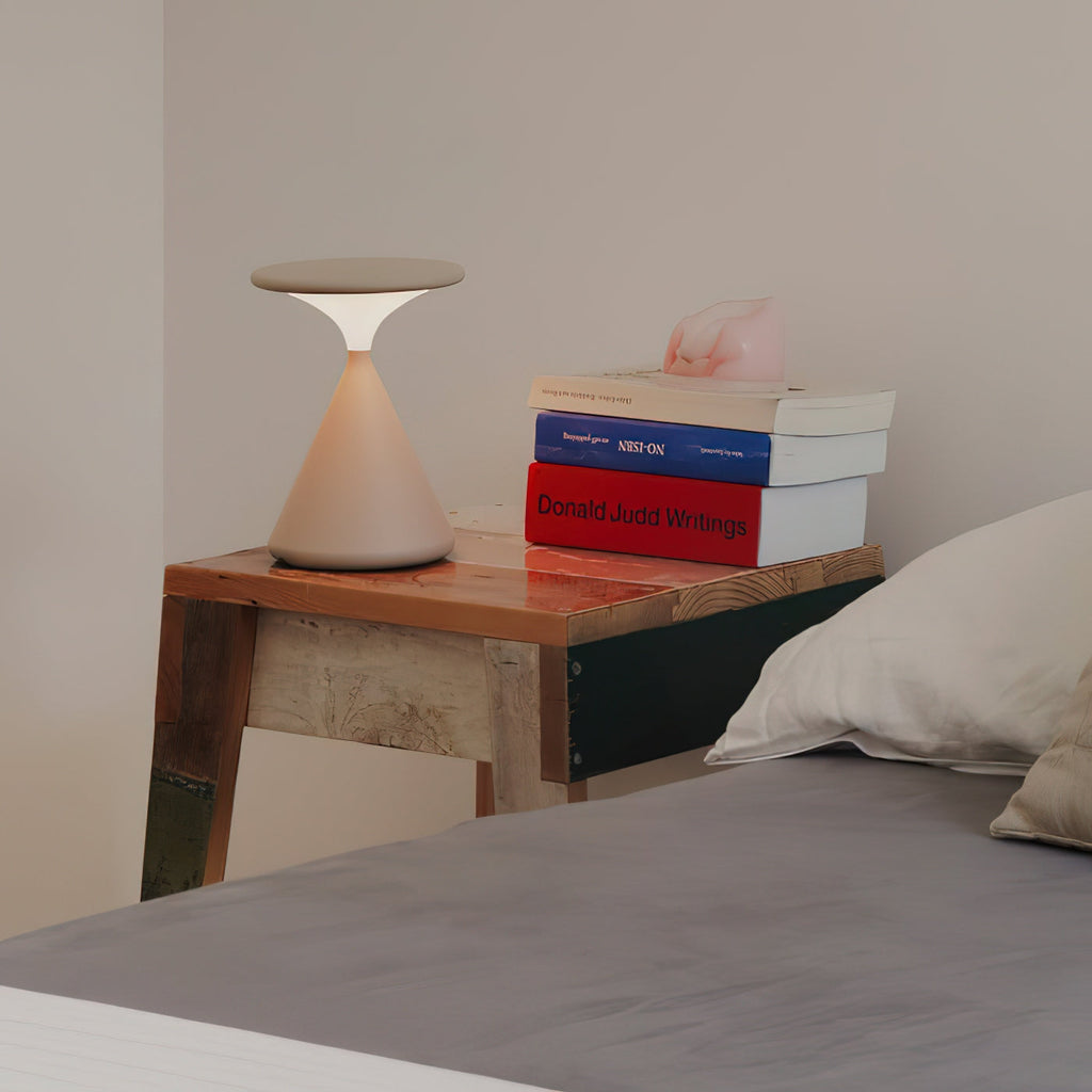 Cream Clérachic table lamp on a rustic wooden bedside table, beside a pile of books and a pillow on a bed.