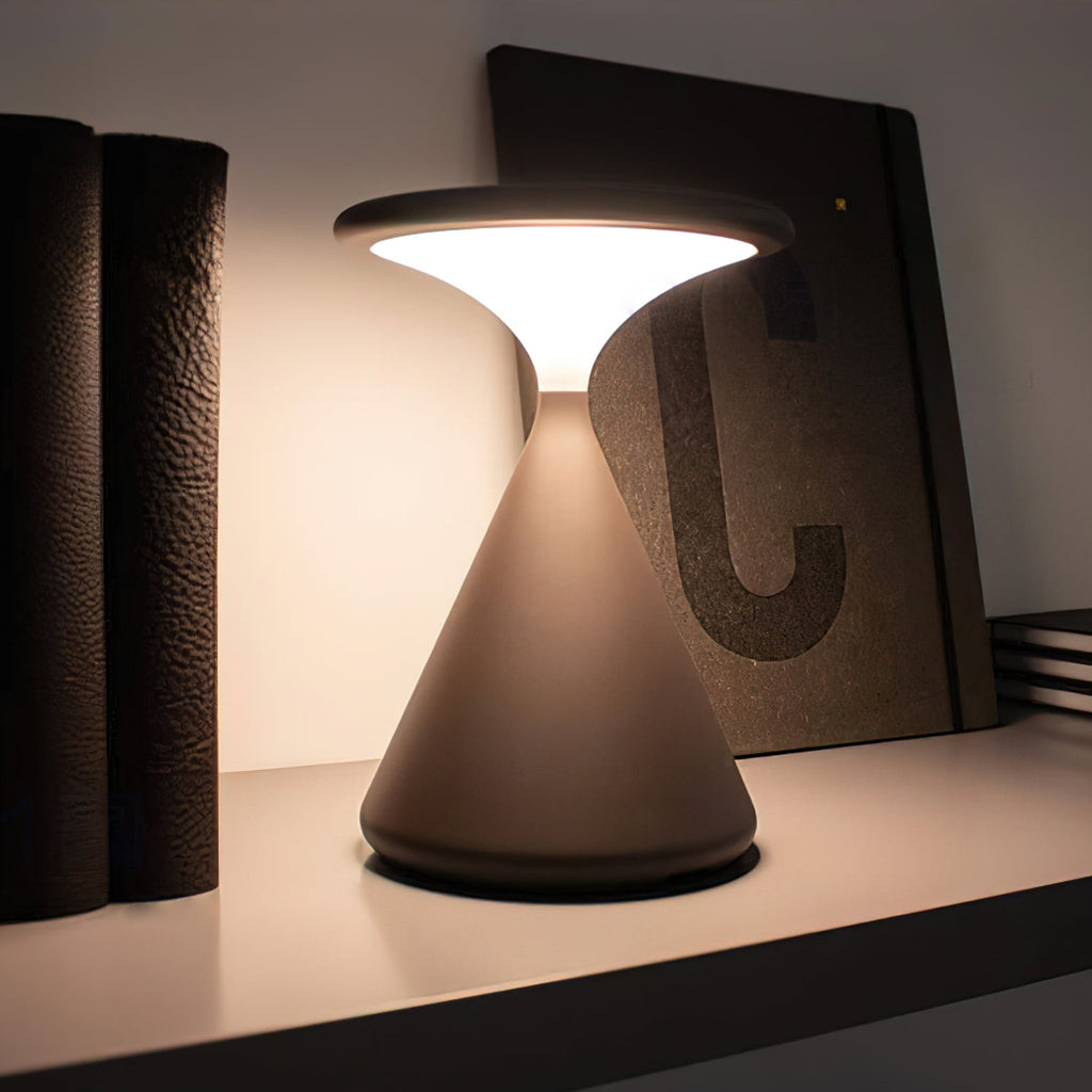 Close view of a Clérachic table lamp lit warm on a shelf, with its disc top and cone base against a dark book.