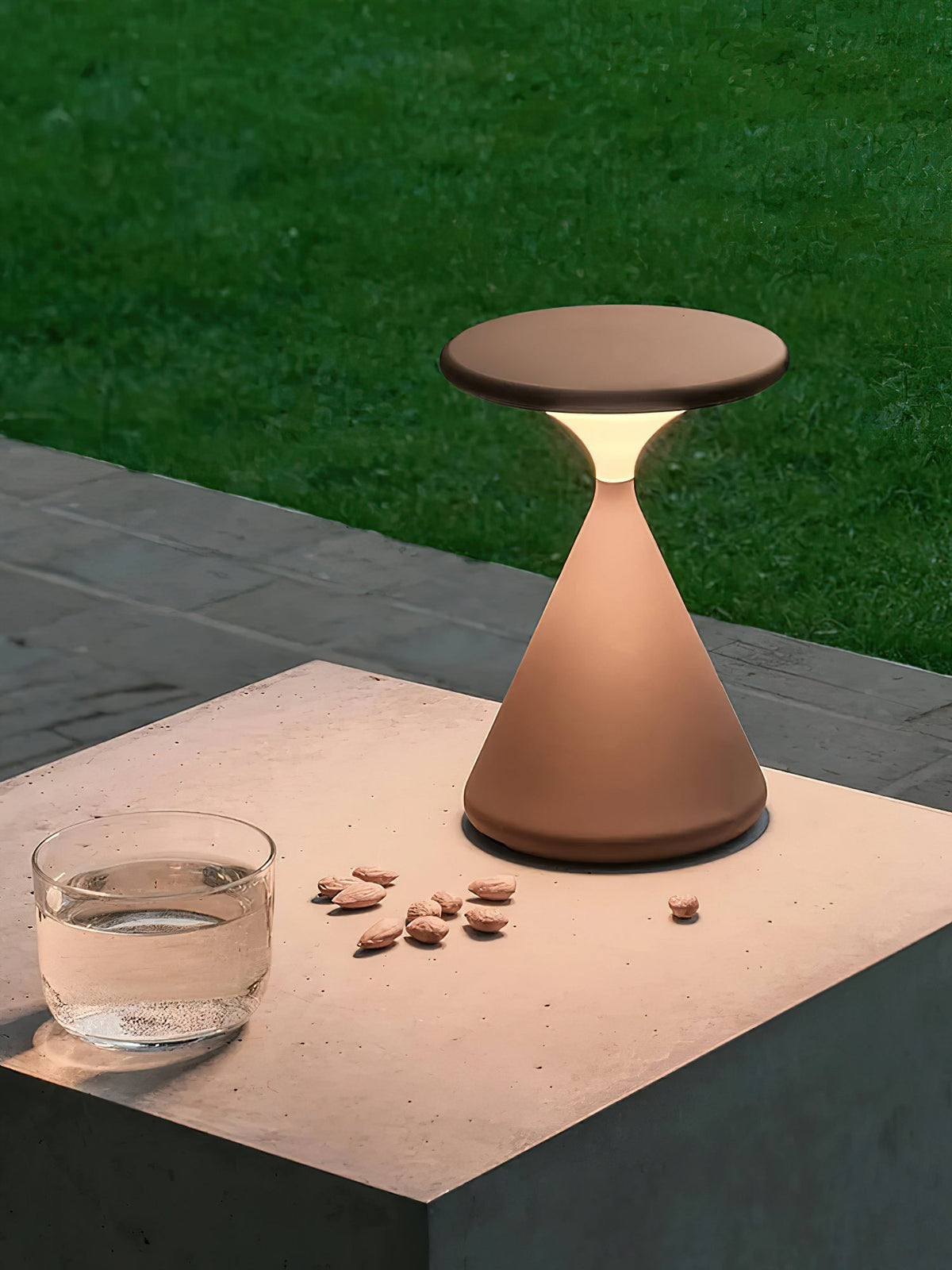 A brown Clérachic table lamp lit on a concrete block outdoors, with a glass of water, a few almonds and grass behind.