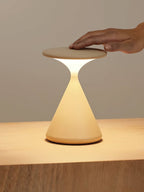 A hand rests on the flat top of this Clérachic table lamp, whose cone base glows warm on a wooden surface.