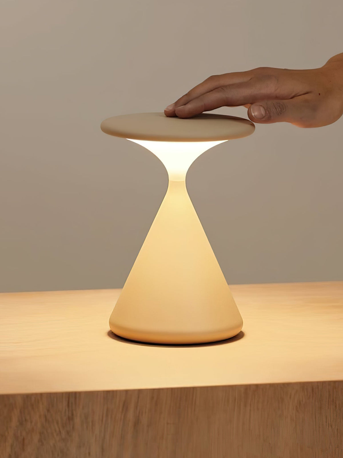 A hand rests on the flat top of this Clérachic table lamp, whose cone base glows warm on a wooden surface.