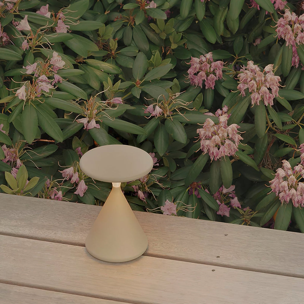 Cream Clérachic table lamp on a pale wooden deck, with pink rhododendron flowers and green leaves behind it.