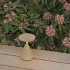 Cream Clérachic table lamp on a pale wooden deck, with pink rhododendron flowers and green leaves behind it.