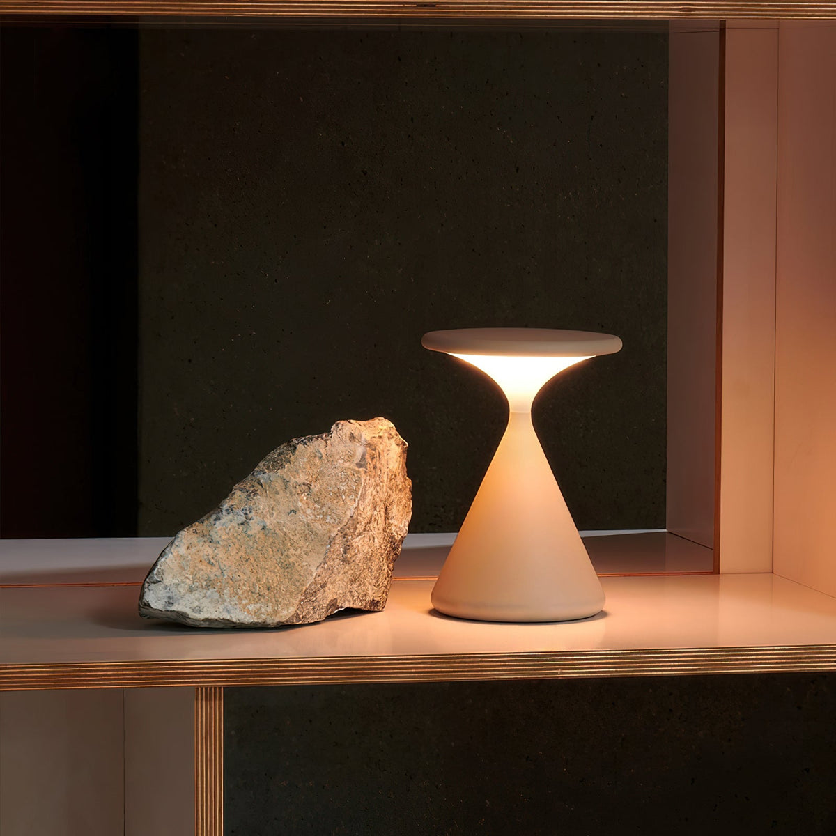 A lit Clérachic table lamp on a wooden shelf beside a rough piece of stone, against a dark wall.