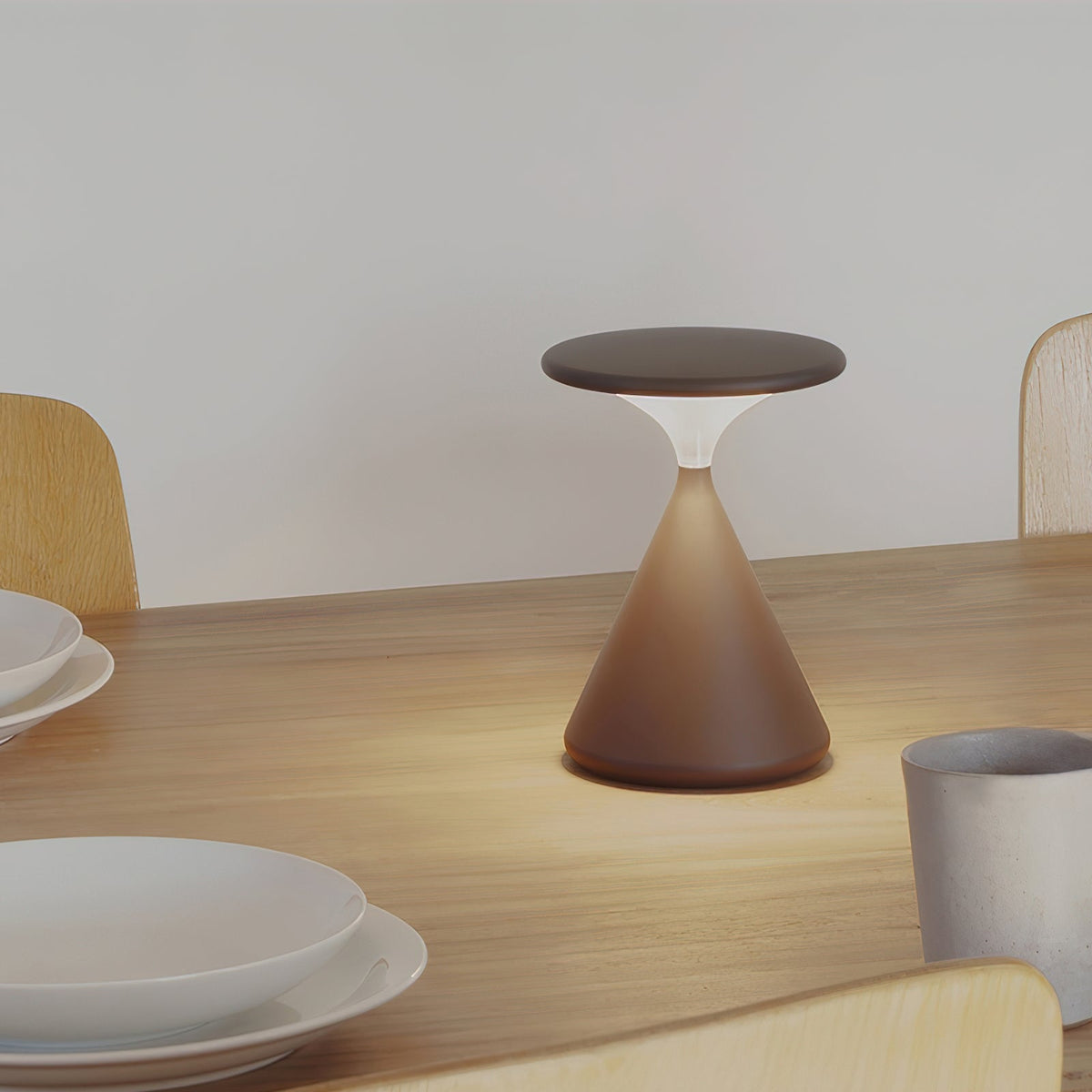 Brown Clérachic table lamp, softly lit, on a wooden dining table with white plates and a grey cup beside it.