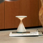 A cream Clérachic table lamp lit on a magazine on the floor, in front of wooden cabinet doors.