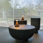 Two small Clérachic table lamps glow on ribbed round side tables in front of a large window onto trees.