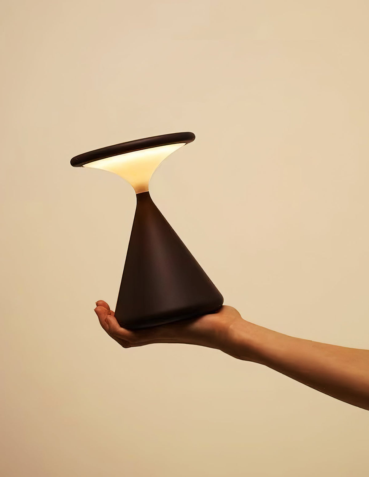 A hand holds a black Clérachic table lamp tilted, its disc top angled to the side as the light glows underneath.