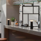 Clérachic cordless table lamps in black spaced along a grey speckled stone bar counter, one set back by a potted herb and one beside a filled martini glass among cocktail tools.