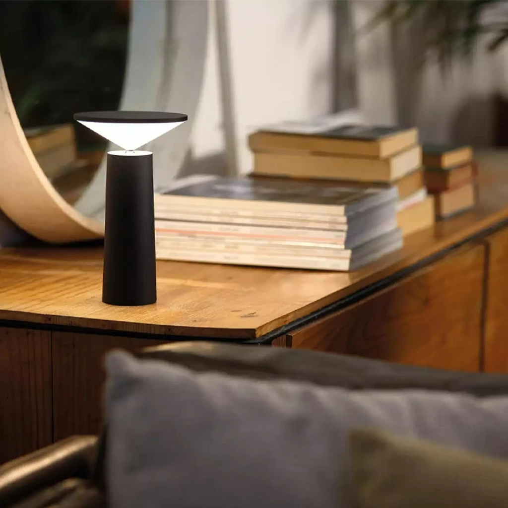 Clérachic cordless table lamp in black on a wood console table, its cone throwing a soft wash of light over stacked books and magazines beside a round mirror.
