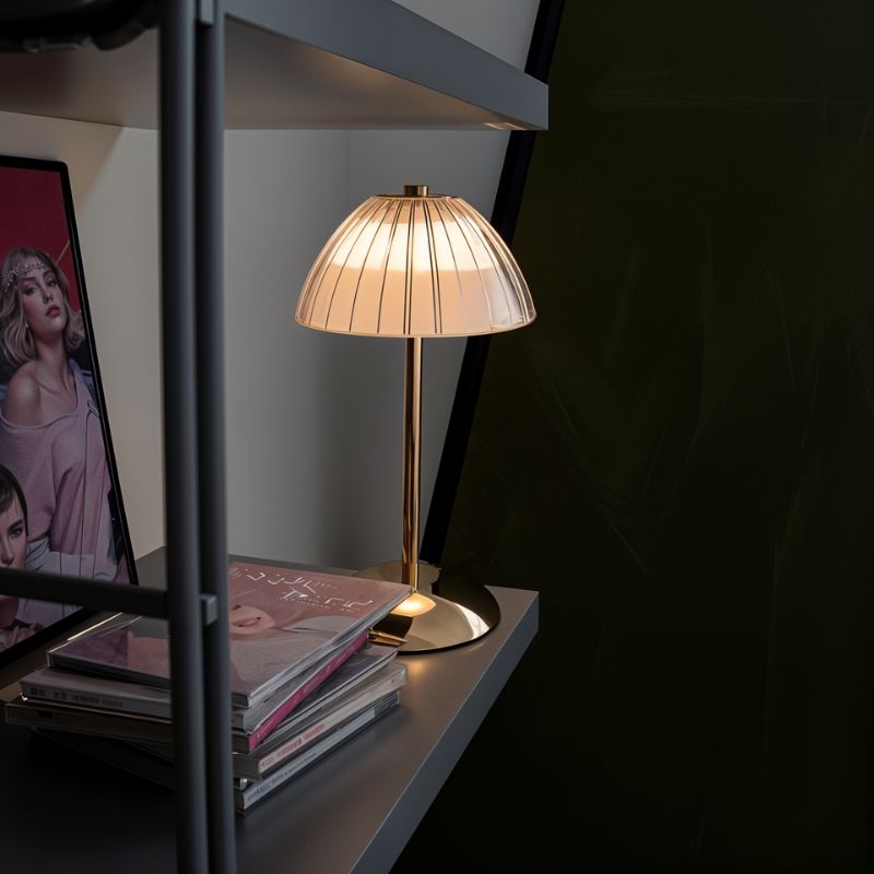 Clérachic cordless table lamp with a ribbed dome shade and slim gold stem, lit on a grey shelf beside stacked magazines.