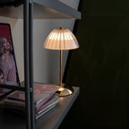 Clérachic cordless table lamp with a ribbed dome shade and slim gold stem, lit on a grey shelf beside stacked magazines.