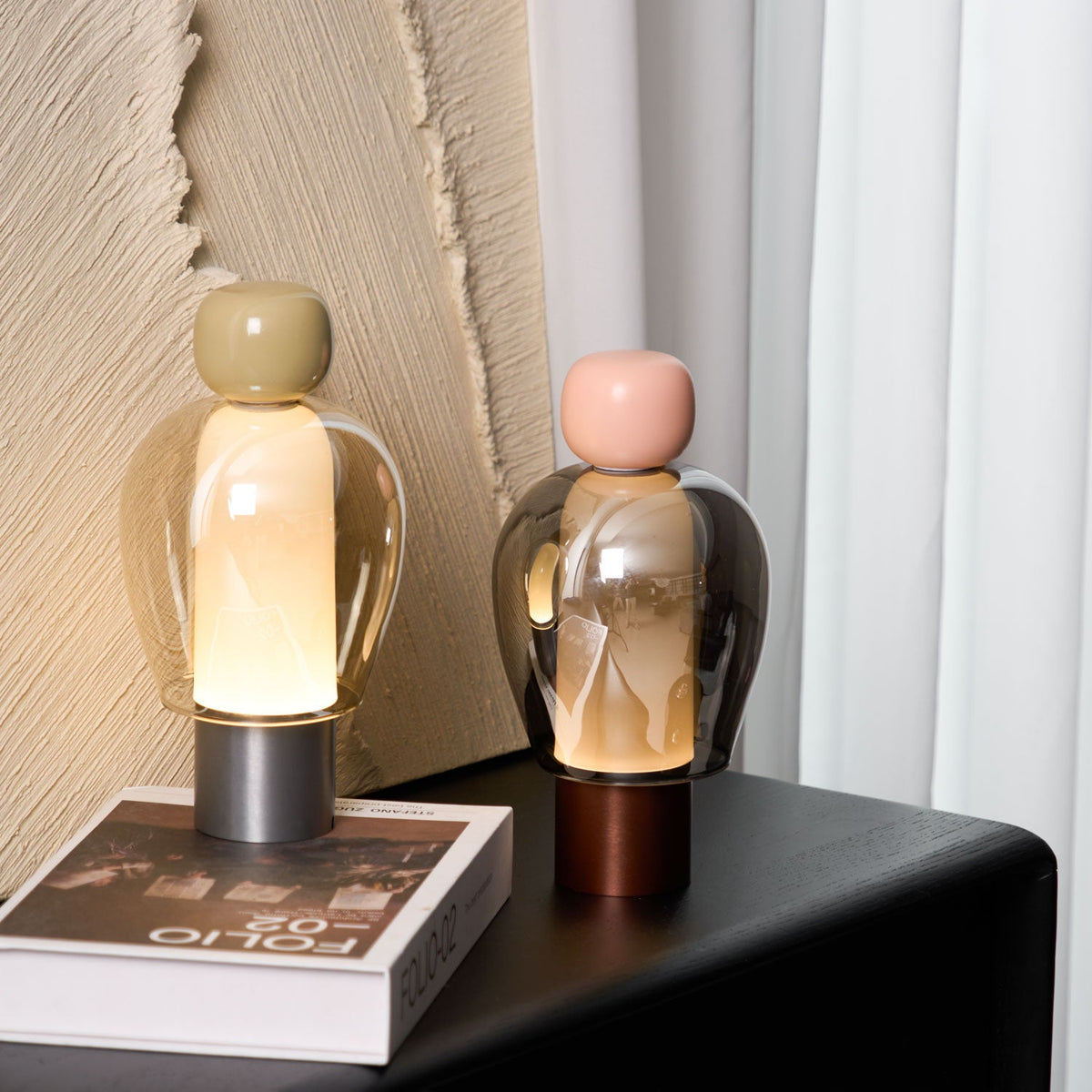 Two lit Clérachic table lamps on a black cabinet, one with an olive cap and one pink, in front of a ridged plaster panel.