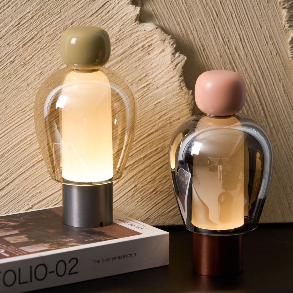 Close view of two lit Clérachic table lamps, their glass domes glowing softly, with an olive cap on the left and pink on the right.