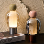 Close view of two lit Clérachic table lamps, their glass domes glowing softly, with an olive cap on the left and pink on the right.