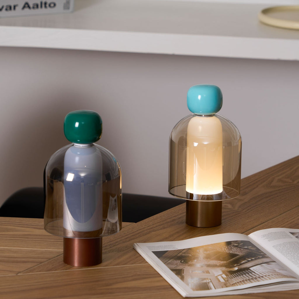 Two Clérachic table lamps on a herringbone wood desk beside an open magazine, one with a green cap and one with a turquoise cap.