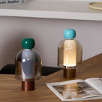 Two Clérachic table lamps on a herringbone wood desk beside an open magazine, one with a green cap and one with a turquoise cap.