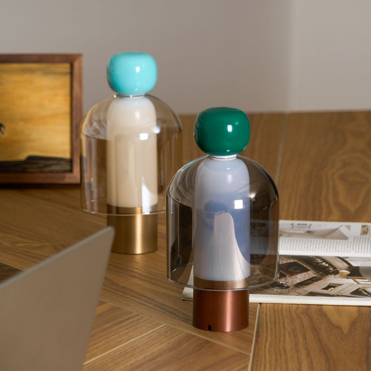 A green-capped and a turquoise-capped Clérachic table lamp on a wood desk, with a framed picture and open magazine behind.