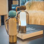 Close pair of Clérachic table lamps on a dark table, a turquoise-capped one in front of a brown-capped lamp lit white-blue.