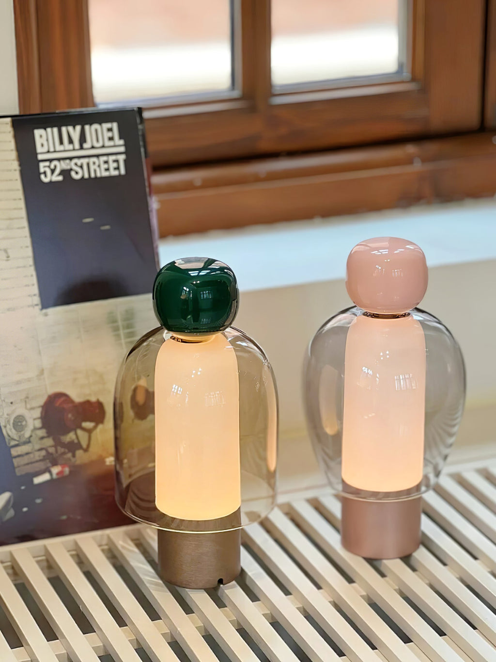 Two lit Clérachic table lamps, a green-capped and a pink-capped one, on a slatted white bench under a small window.