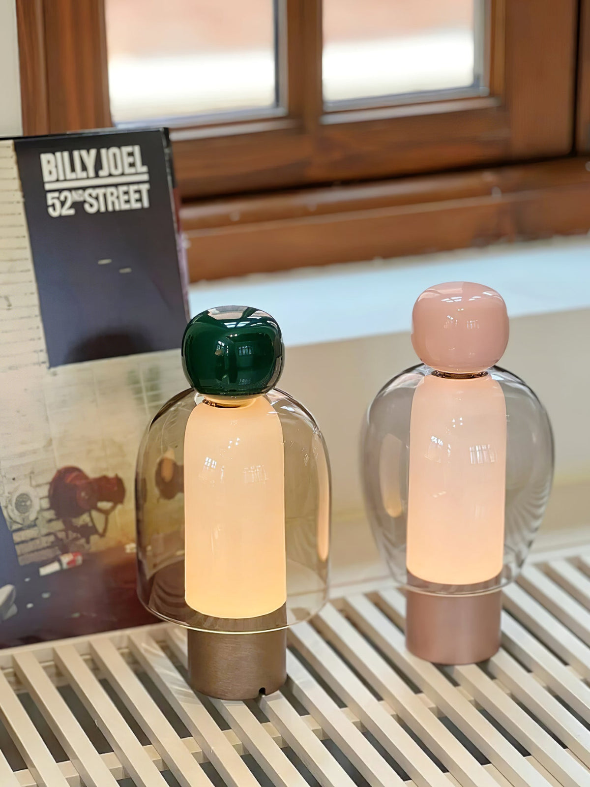Two lit Clérachic table lamps, a green-capped and a pink-capped one, on a slatted white bench under a small window.