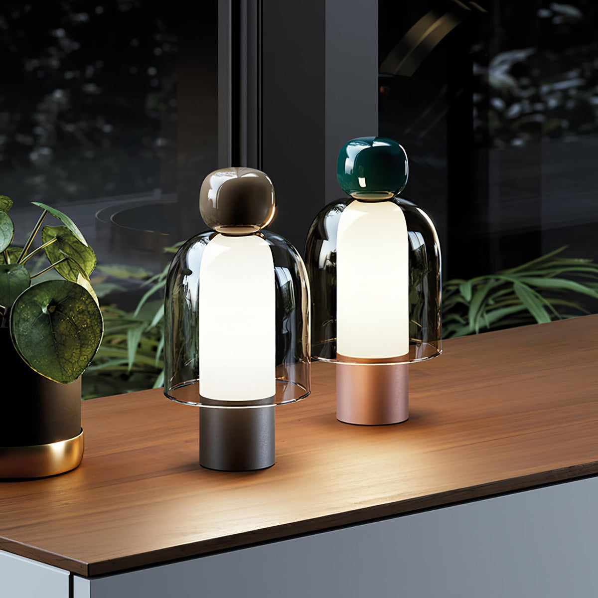 Two lit Clérachic table lamps on a walnut-veneer sideboard by a window, with a leafy plant at the left and a dark garden outside.