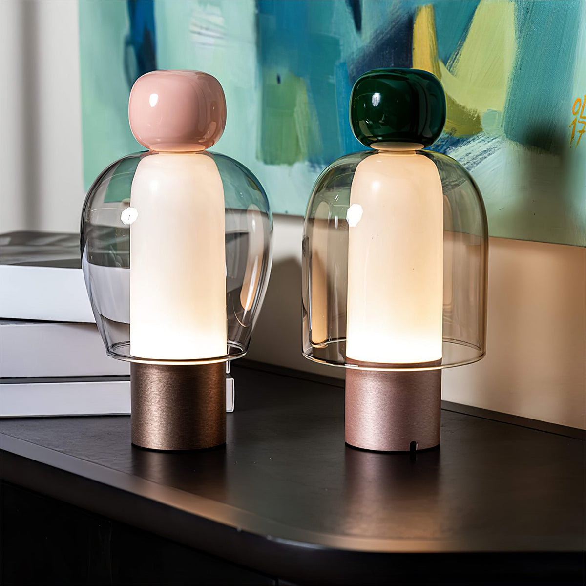 Two lit Clérachic table lamps, one with a pink cap and one dark green, on a black ledge in front of a blue and yellow painting.