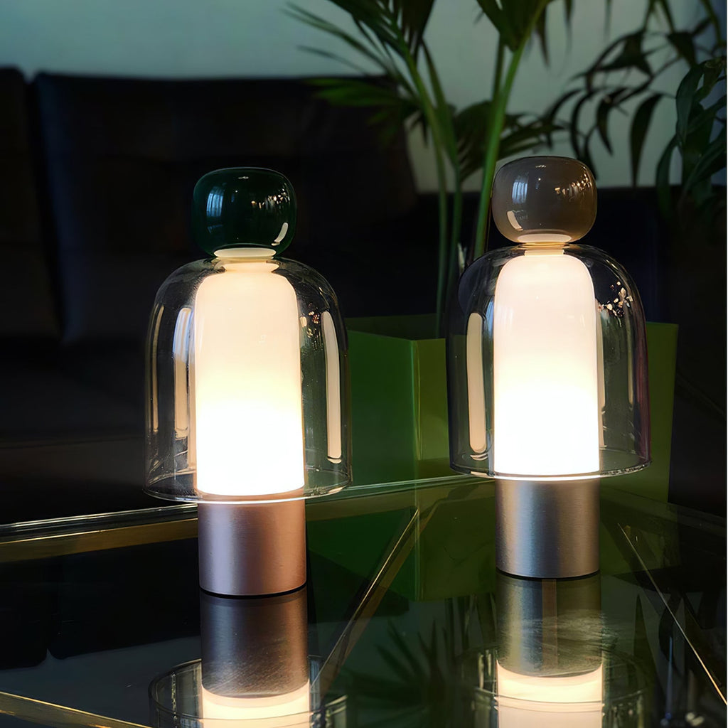 Two lit Clérachic table lamps with dark green and brown caps on a glass shelf, their bases reflected in the surface below.
