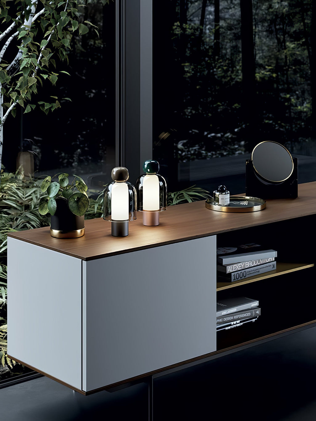 Wide view of a sideboard with two small lit Clérachic table lamps on top, beside a plant, a tray and a round mirror.