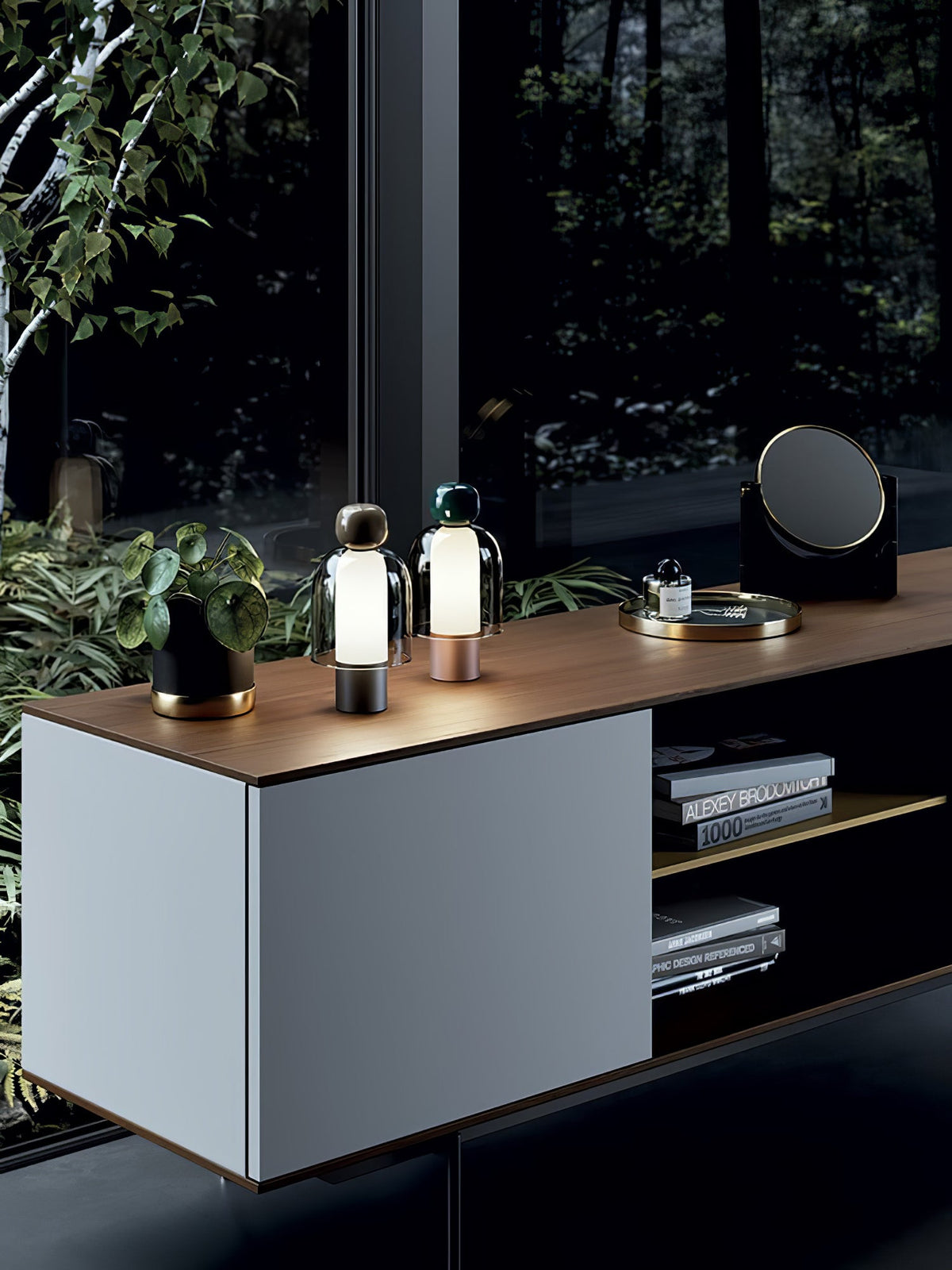 Wide view of a sideboard with two small lit Clérachic table lamps on top, beside a plant, a tray and a round mirror.