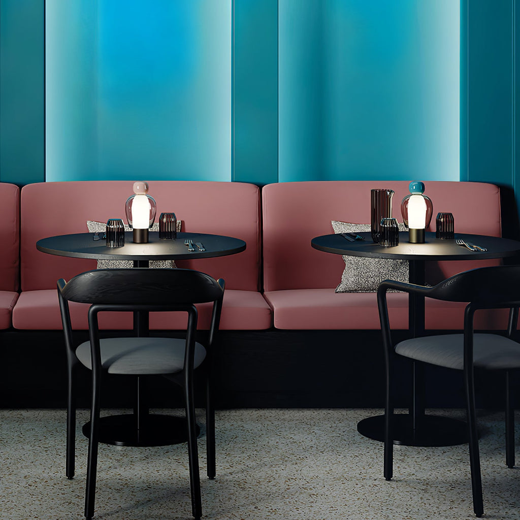 Small Clérachic table lamps glowing on café tables, in front of a rose-pink banquette and a teal wall with black chairs.