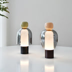 Two lit Clérachic table lamps on a white round table, one with an olive cap and one with a peach cap, against a pale wall.
