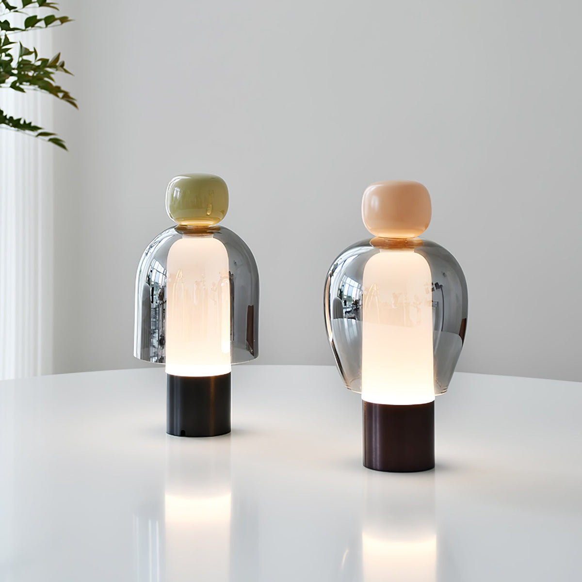 Two lit Clérachic table lamps on a white round table, one with an olive cap and one with a peach cap, against a pale wall.